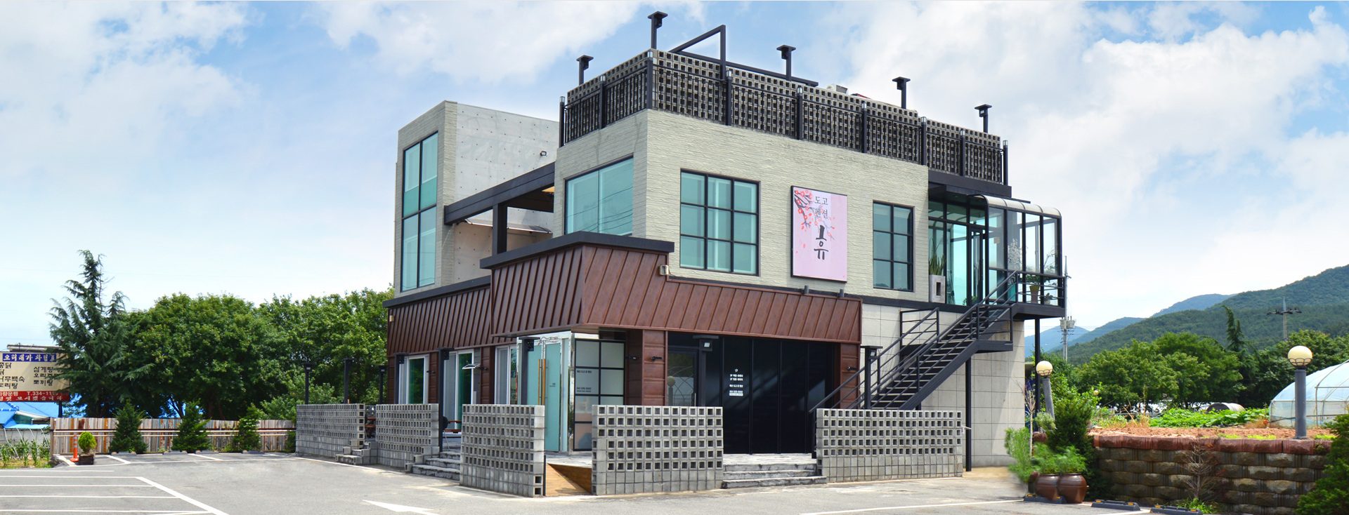 맑은 날 도고휴펜션 건물 외관 — 갈색 금속 외벽·철제 계단·옥상 난간, 뒤로 산